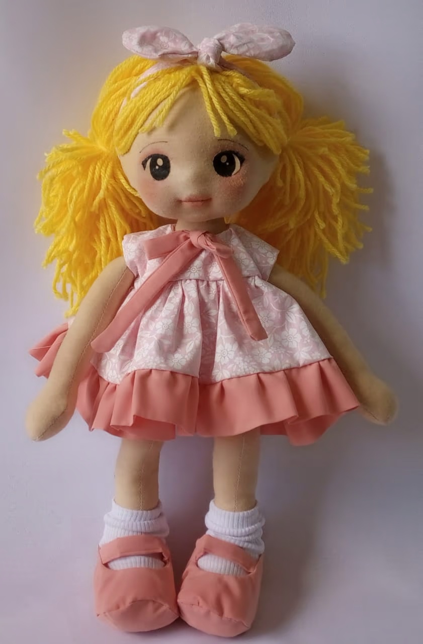 Boneca de pano loira com vestido rosa claro, sapatos rosa escuro e olhos escuros.