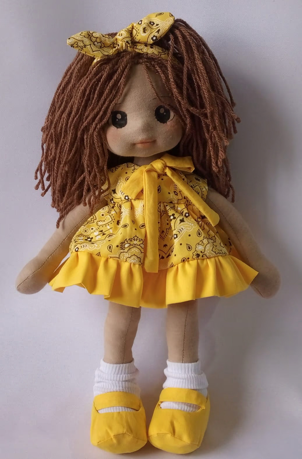 Boneca de pano morena com vestido amarelo estampado, sapatos amarelos e cabelo castanho.